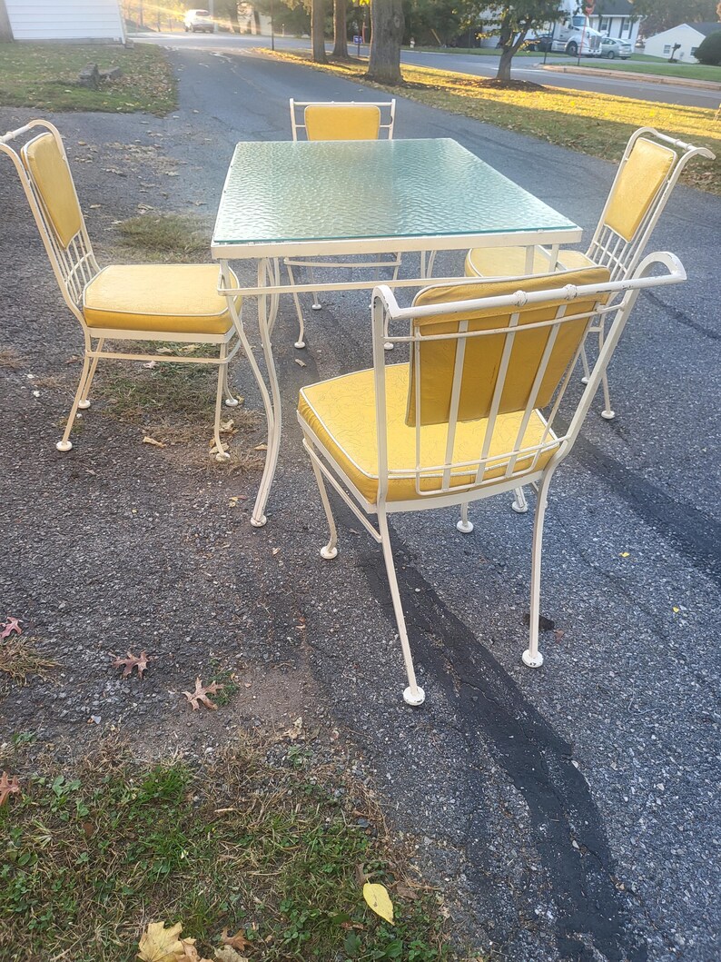 May include: A white metal patio dining set with a glass table top and four chairs. The chairs have yellow cushions.