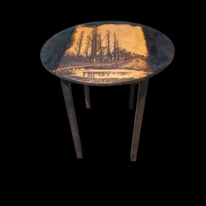 May include: A round wooden table with a black finish and a wood top. The top has a burnt-in image of a landscape with trees and a river.