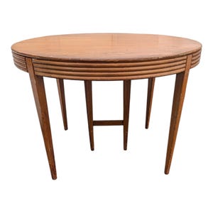 May include: A round, light brown wooden table with four angled legs. The table's edge features a series of horizontal, grooved lines. The table is a vintage piece, perfect for a living room or dining area.