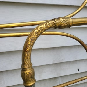 Op de afbeelding: Detailopname van een vintage messing bedframe. Het frame heeft sierlijke bloemendetails op het gebogen hoofdbord en verbindingsstukken. Het messing heeft een warme, gouden tint, met enkele plekken die lichte patina vertonen.