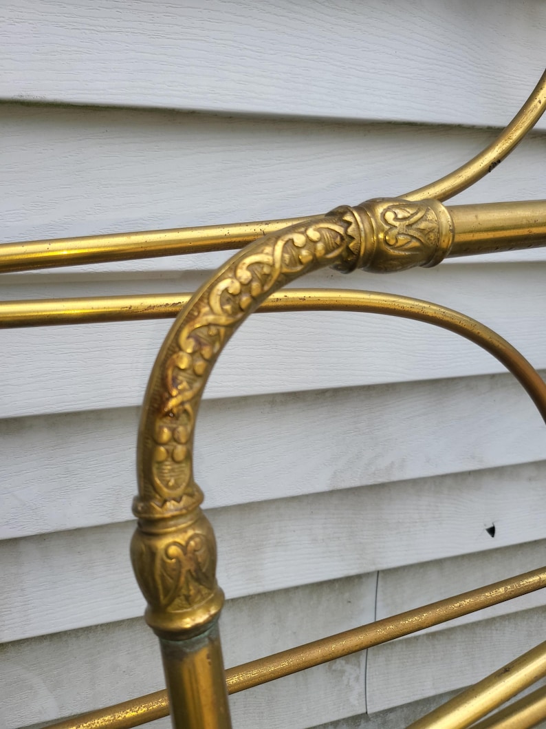 Op de afbeelding: Detailopname van een vintage messing bedframe. Het frame heeft sierlijke bloemendetails op het gebogen hoofdbord en verbindingsstukken. Het messing heeft een warme, gouden tint, met enkele plekken die lichte patina vertonen.