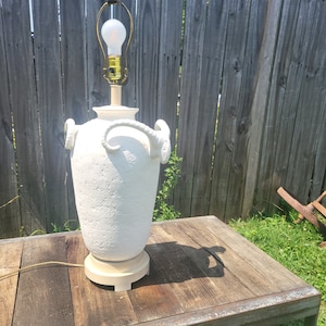 May include: White ceramic table lamp with ram's horns and a textured finish. The lamp has a white light bulb and a gold-colored base.