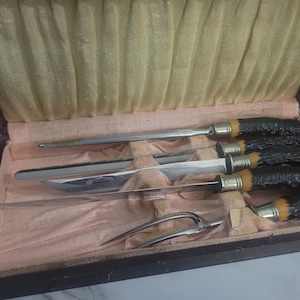 May include: A vintage carving set in a wooden case. The set includes a carving knife, a sharpening steel, and a carving fork. The handles are dark brown with a yellow accent. The case has a pink interior.