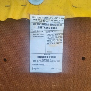 May include: A close-up of a brown fabric tag with white text. The tag reads "Under penalty of law this tag not to be removed except by the consumer. All new material consisting of urethane foam. Lic. No. N.Y. 9302 (N.C.) Space for stamp (when required) Certification is made by the manufacturer that the materials in this article are described in accordance with law. Made by Carolina Forge Division of Lee L. Woodard Sons, Inc. Date of Delivery. Frame No. 5 Color M-14730"