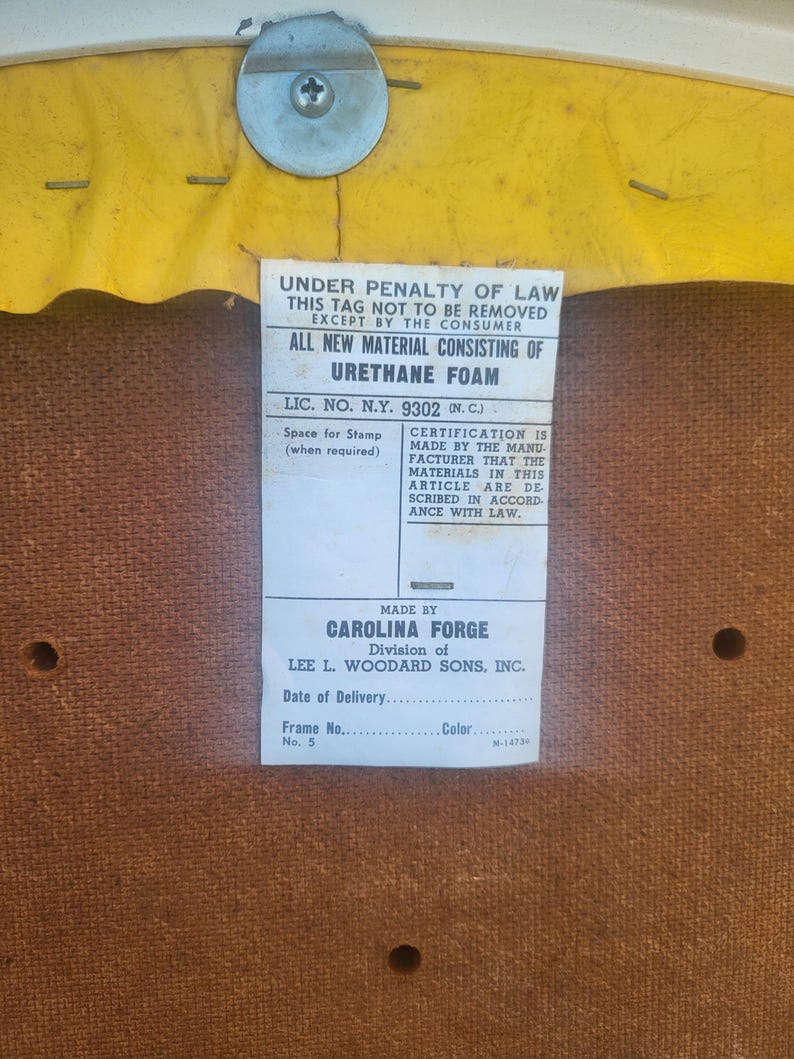 May include: A close-up of a brown fabric tag with white text. The tag reads "Under penalty of law this tag not to be removed except by the consumer. All new material consisting of urethane foam. Lic. No. N.Y. 9302 (N.C.) Space for stamp (when required) Certification is made by the manufacturer that the materials in this article are described in accordance with law. Made by Carolina Forge Division of Lee L. Woodard Sons, Inc. Date of Delivery. Frame No. 5 Color M-14730"