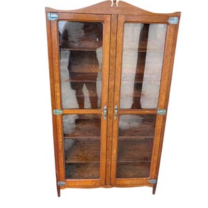 May include: A tall, rectangular wooden cabinet with two glass-paneled doors. The cabinet features a medium brown wood grain finish, silver-toned metal hinges at the corners, and vertical metal handles. The top has a decorative arched peak.