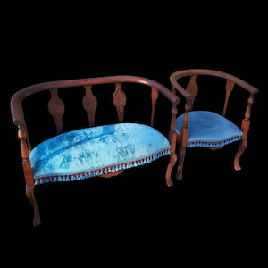 May include: Two antique wooden chairs with curved backs and blue velvet seats. The chairs have ornate carved details on the backrests and legs. The seats are trimmed with a decorative fringe.