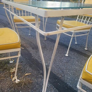 May include: White metal patio table with a glass top and four chairs with yellow cushions. The table and chairs are in a backyard setting.