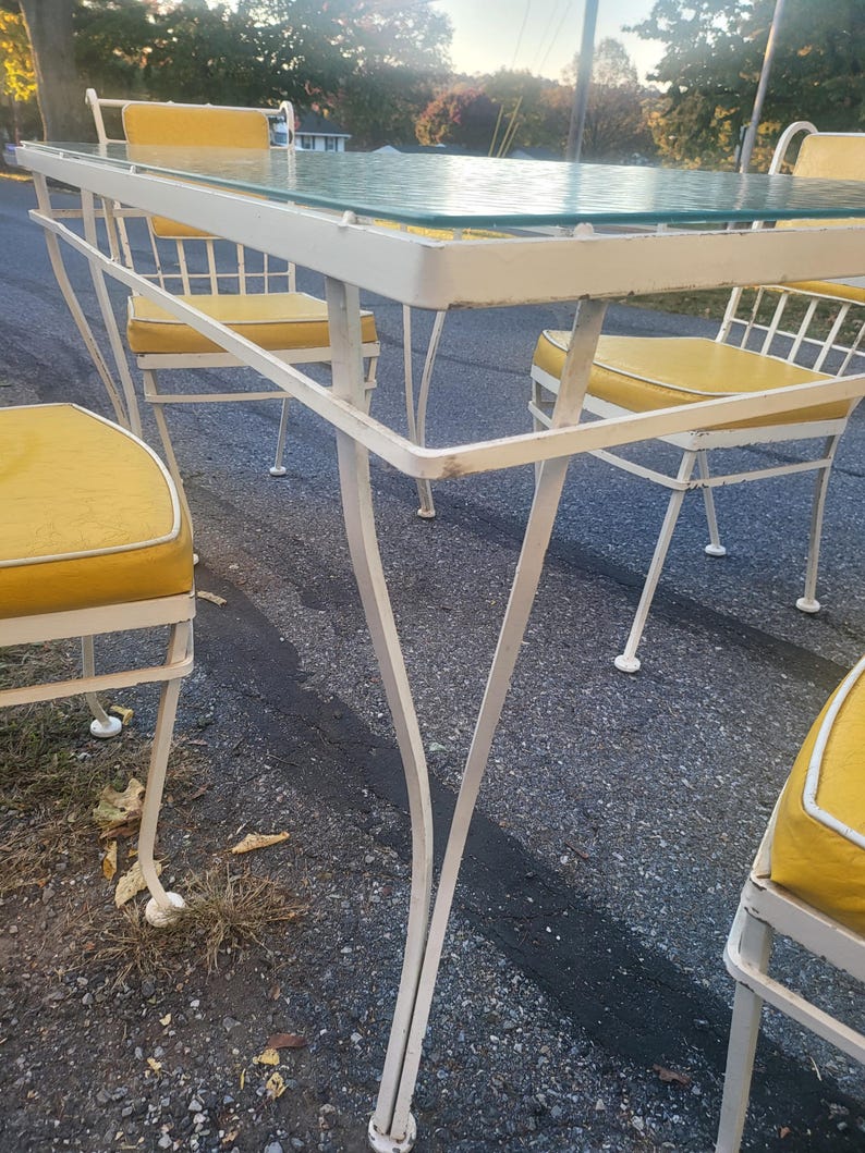 May include: White metal patio table with a glass top and four chairs with yellow cushions. The table and chairs are in a backyard setting.