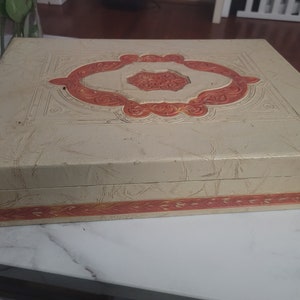 May include: A vintage, cream-colored box with a raised, orange and white design. The box has a decorative border around the edge.