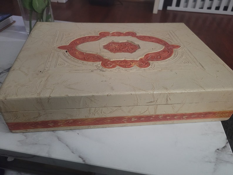 May include: A vintage, cream-colored box with a raised, orange and white design. The box has a decorative border around the edge.