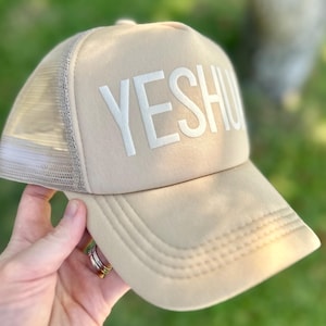 Könnte beinhalten: Beigefarbene Trucker-Cap mit dem Wort "YESHU" in weißen Blockbuchstaben auf der Vorderseite. Die Kappe hat eine Netzrückseite und einen gebogenen Schirm. Die Kappe wird von einer Hand gehalten.