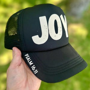 Puede incluir: Gorra de camionero negra con la palabra "JOY" en letras blancas grandes y desgastadas en la parte delantera. El texto "PSALM 16:11" está impreso en blanco en el lateral. La gorra tiene una parte trasera de malla y una visera curva.