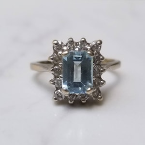 May include: A silver ring with a light blue aquamarine gemstone surrounded by a halo of small diamonds.