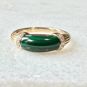 May include: A gold ring featuring a horizontal, rectangular malachite gemstone with a deep green banded pattern. The band is ribbed on both sides, accented by small, clear, round-cut stones set into the metal where the malachite meets the band.