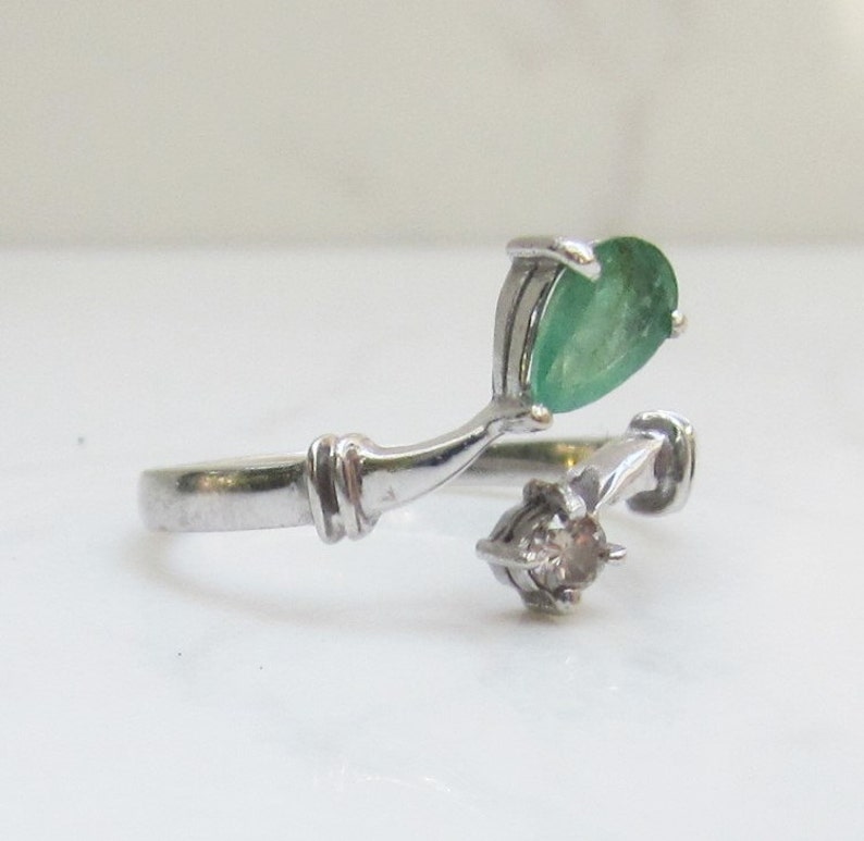 Puede incluir: Un anillo de plata con una esmeralda de forma de pera y un peque&ntilde;o diamante redondo. El anillo tiene un dise&ntilde;o &uacute;nico con la esmeralda engastada en una banda curva.