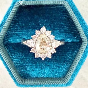 May include: A silver-toned ring featuring a central pear-shaped champagne-colored gemstone surrounded by a halo of small round white stones and two larger pear-shaped side stones, all set within a teal velvet hexagonal ring box.