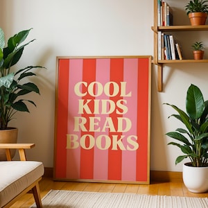 Könnte beinhalten: Gerahmtes Poster mit dem Text "COOL KIDS READ BOOKS" in goldfarbenen Buchstaben auf einem rot-rosa gestreiften Hintergrund. Das Poster wird in einem Raum mit Pflanzen und einem Holzstuhl ausgestellt.