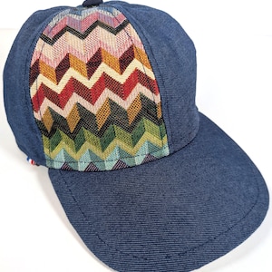 Puede incluir: Una gorra de béisbol con tela azul oscuro tipo mezclilla en la visera y los paneles traseros, contrastada por un panel frontal con un vibrante patrón de chevrón multicolor en tonos rojo, naranja, amarillo, verde y azul claro.