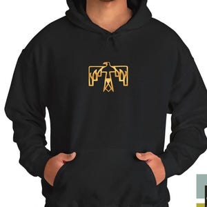 Puede incluir: Una sudadera con capucha negra que presenta un gráfico de pájaro geométrico de color dorado centrado en el pecho. La sudadera incluye un bolsillo tipo canguro frontal, mangas largas y capucha con cordón, con un diseño funcional.