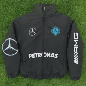 May include: A black zip-up jacket featuring the Mercedes-Benz logo on the chest and left sleeve, with "PETRONAS" printed across the front. The right sleeve displays "AMG" in white. The jacket is on a green background.