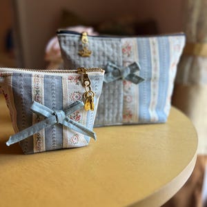 Peut inclure: Deux pochettes en tissu avec un motif à rayures bleu clair et crème agrémenté de petites fleurs. Chaque pochette possède une fermeture éclair dorée avec une breloque décorative, une texture matelassée et un nœud en tissu bleu clair.