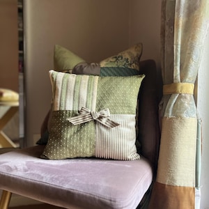 May include: A square scatter cushion featuring a patchwork design of sage green and cream fabrics. The front displays alternating panels of striped and dotted textures, accented by a small fabric bow tied in the centre. The cushion is placed on a chair.