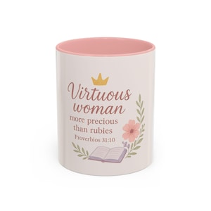 May include: A white ceramic mug with a pink interior. The mug features the text "Virtuous woman more precious than rubies Proverbs 31:10" with a gold crown, floral, and book illustrations. The mug is designed for beverages.