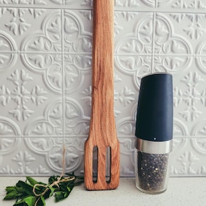 May include: A wooden cooking utensil with a slotted design, a black and silver pepper grinder filled with peppercorns, and a bundle of fresh mint on a white countertop. The background features a white, patterned wall.
