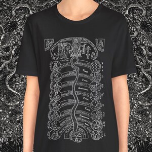 Könnte beinhalten: Schwarzes T-Shirt mit detailliertem weißem Grafikdruck. Das Design zeigt ein zentrales vertikales Element mit Text und Illustrationen, flankiert von dekorativen Bordüren. Das T-Shirt wird von einer Person vor einem schwarz-weiß gemusterten Hintergrund getragen.