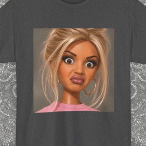 Könnte beinhalten: Ein anthrazitfarbenes Kurzarm-T-Shirt mit einer zentrierten quadratischen Grafik einer blonden Puppe mit großen Augen, Creolen und einem rosa Oberteil. In der unteren rechten Ecke befindet sich ein schwarzes Logo mit dem Text BR BLACKASH RECORDS.