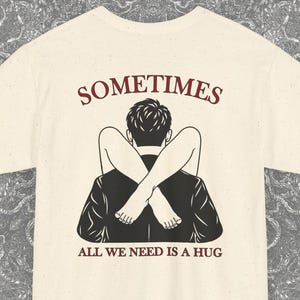 Könnte beinhalten: Ein cremefarbenes Kurzarm-T-Shirt mit einer schwarzen grafischen Illustration einer Person, die von hinten umarmt wird, wobei die Beine um die Schultern geschlungen sind. Der Text SOMETIMES steht darüber und ALL WE NEED IS A HUG darunter in Rot.