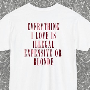 Può includere: T-shirt bianco con la scritta bordeaux "EVERYTHING I LOVE IS ILLEGAL EXPENSIVE OR BLONDE". La maglietta ha maniche corte e girocollo. Il logo Blackash Records si trova in basso a destra.