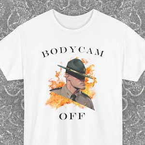Op de afbeelding: Wit T-shirt met de tekst "BODYCAM OFF" boven en onder een illustratie van een man met een rangerhoed, omringd door vlammen. Het shirt heeft een ronde hals en korte mouwen. Het Blackash Records-logo staat rechtsonder.
