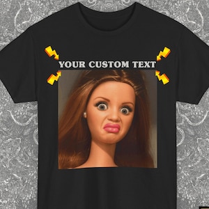 Può includere: Una t-shirt nera a maniche corte con una grafica rettangolare centrale raffigurante una bambola con lunghi capelli castani e un'espressione sorpresa. Sopra la bambola, il testo YOUR CUSTOM TEXT è affiancato da quattro icone di fulmini gialli.