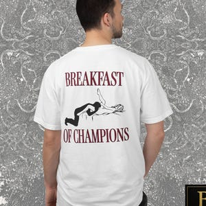 Pode incluir: Camiseta branca com o texto "BREAKFAST OF CHAMPIONS" em bordô. A camiseta apresenta um gráfico em preto e branco de duas figuras. O logotipo da Blackash Records está no canto inferior direito.