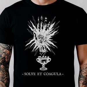 Puede incluir: Camiseta negra con un gráfico blanco detallado de una mano, un cáliz y otros símbolos con líneas radiantes. El texto "- SOLVE ET COAGULA -" está debajo. La camiseta tiene cuello redondo y mangas cortas.
