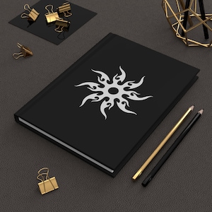 Puede incluir: Un diario negro con un diseño de sol blanco en la portada. El diario está rodeado de lápices dorados y negros, clips dorados y un cuadrado negro con más clips dorados. El diario está sobre una superficie gris oscuro.