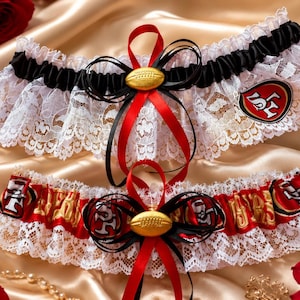 Könnte beinhalten: Zwei Hochzeitsstrumpfbänder aus weißer Spitze, schwarzen und roten Satinbändern sowie einem goldenen Football-Anhänger. Ein Strumpfband hat ein San Francisco 49ers-Logo-Aufnäher, das andere ein wiederkehrendes Team-Logo-Muster auf rotem Satin.