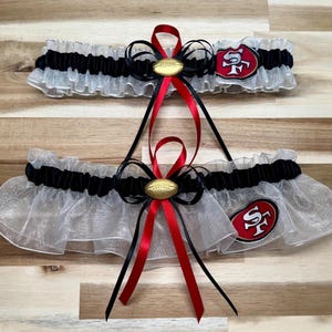 May include: Two ruffled garters, one above the other, featuring black and white fabric with red and black ribbons. Each garter has a gold football-shaped embellishment and a red and gold team logo patch.