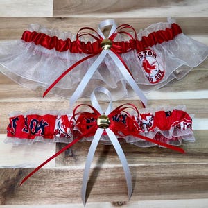 Puede incluir: Dos ligas de boda. La liga superior es blanca con ribete rojo y un lazo rojo y blanco. La liga inferior es roja con detalles blancos y un lazo rojo y blanco. Ambas tienen un tema de béisbol.