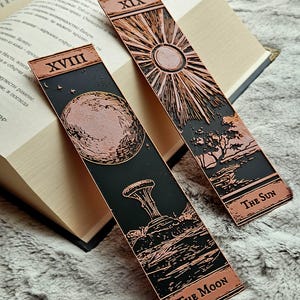 Puede incluir: Dos marcapáginas metálicos con diseños color cobre y fondos negros. Uno presenta el sol con el texto "The Sun", y el otro la luna con el texto "The Moon". Ambos tienen números romanos en la parte superior y están sobre un libro abierto.