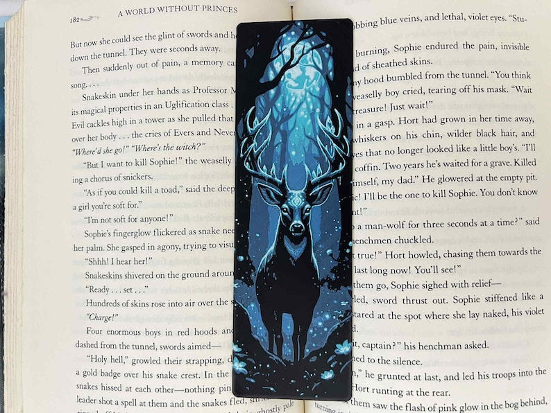 May include: A rectangular bookmark featuring a stylized illustration of a stag in a forest. The stag is black with large antlers, set against a backdrop of a blue and teal forest scene. The bookmark is placed on a book.