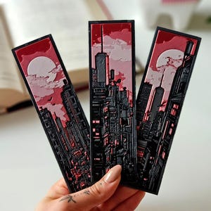 Peut inclure: Trois marque-pages noirs et rouges représentant un paysage urbain. Chaque marque-page présente une illustration détaillée d'une ligne d'horizon urbaine avec un ciel rouge, une pleine lune et des nuages. Les marque-pages sont tenus dans une main.
