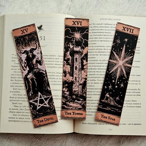Puede incluir: Tres marcadores de cartas del tarot con fondos negros e ilustraciones rosas. El Diablo, La Torre y La Estrella se muestran en un libro abierto. Cada marcador tiene un diseño diferente y el texto del nombre de la carta.