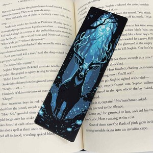 May include: Black bookmark featuring a majestic stag with large antlers, set against a blue and teal forest scene. The bookmark is rectangular and is placed on an open book.