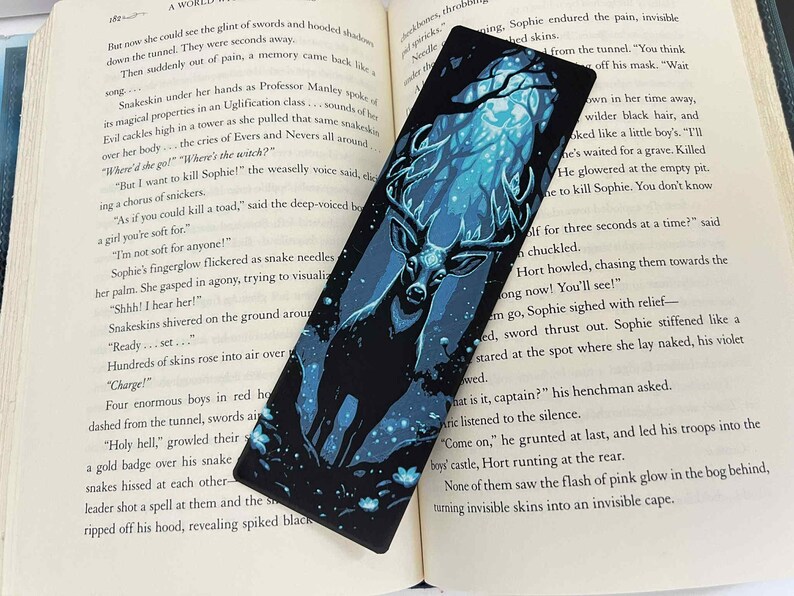 May include: Black bookmark featuring a majestic stag with large antlers, set against a blue and teal forest scene. The bookmark is rectangular and is placed on an open book.