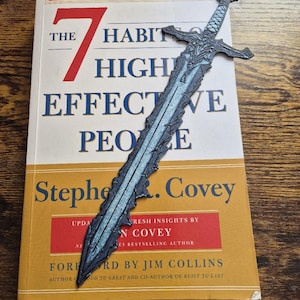 Könnte beinhalten: Ein dekorativer schwertförmiger Lesezeichen liegt auf einem Buch mit dem Titel "The 7 Habits of Highly Effective People" von Stephen Covey. Das Lesezeichen ist grau und blau, und das Buch hat einen gelben Einband mit rotem und schwarzem Text.