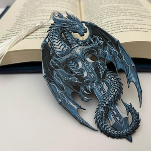Peut inclure: Marque-page en forme de dragon bleu détaillé avec un pompon argenté, posé sur un livre ouvert. Le dragon est finement conçu, tenant une épée avec des runes. Les ailes sont déployées, et les écailles du dragon sont visibles.