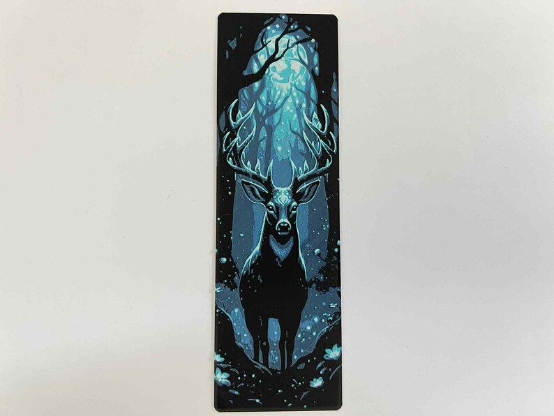 May include: A rectangular bookmark featuring a stylized illustration of a stag in a forest. The stag is black with large antlers, set against a backdrop of a blue and teal forest scene. The bookmark is approximately 2 inches wide and 6 inches tall.